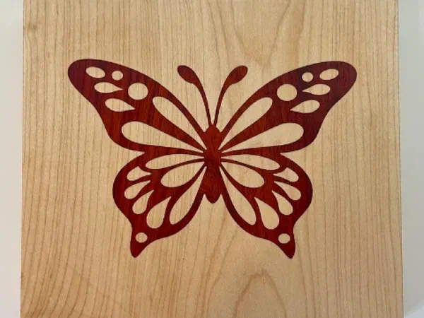 A red padauk butterfly inlaid flush into a maple panel, cut as a V-carve inlay.