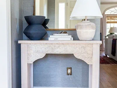 A pale carved console table against a wall with a lamp and vases on top.