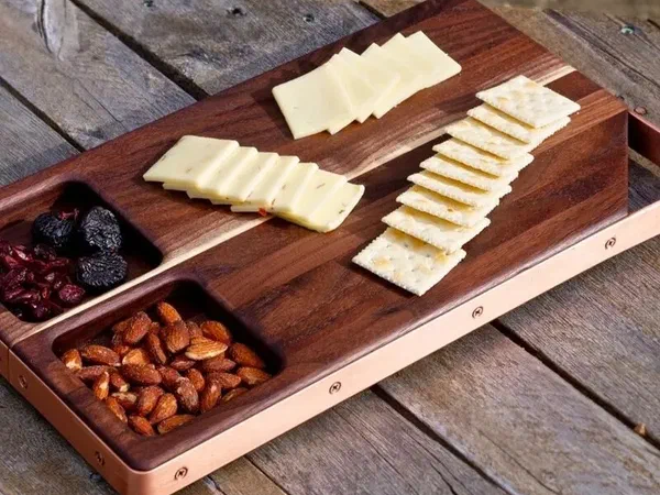 A walnut serving board with cheese, crackers, nuts and fruit arranged on it.